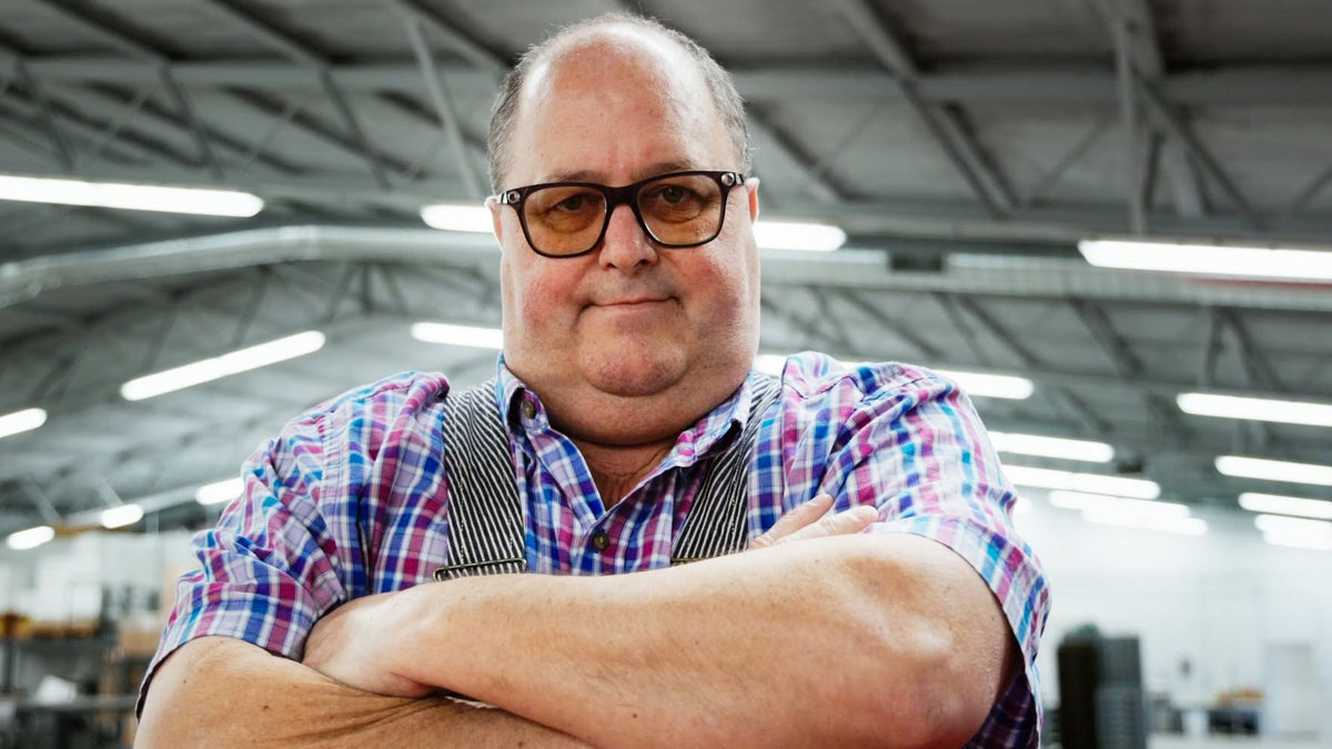 A man with glasses and a receding hairline stands with arms crossed in a bright industrial warehouse setting, wearing a blue, purple, and white plaid button-down shirt with a dark striped apron over his chest.